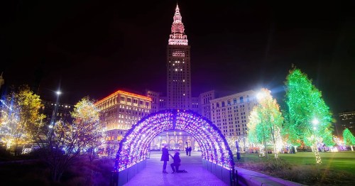 A colorful light display at night with a tall building in the background and a couple walking under a lit arch.