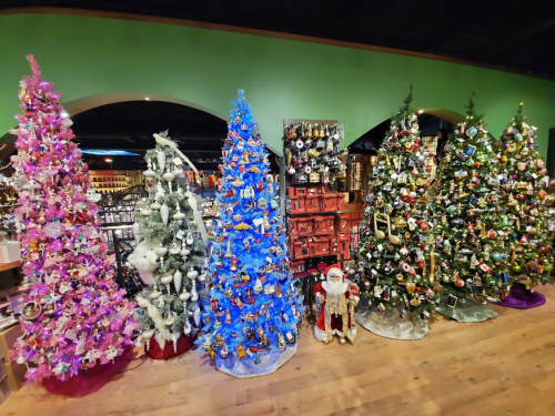 A festive display of five decorated Christmas trees in various colors, with ornaments and a Santa figure nearby.