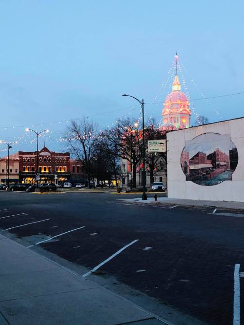 A street view at dusk featuring a lit building and festive lights, with a mural on a wall and parked cars.