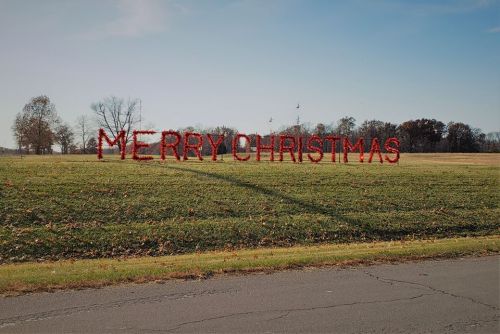 A grassy field with a large "MERRY CHRISTMAS" sign made of red letters against a clear blue sky.