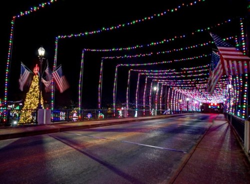 A brightly lit bridge adorned with colorful lights and American flags, with a festive tree in the background at night.