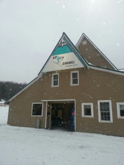 A snow-covered building with a sign reading "Sawmill" and a mountain logo, set in a winter landscape.