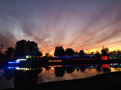 Colorful lights reflect on a calm lake at sunset, with a vibrant sky and silhouetted trees in the background.