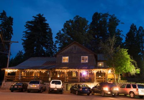 A rustic wooden building with string lights, surrounded by trees, and parked cars in the foreground at dusk.