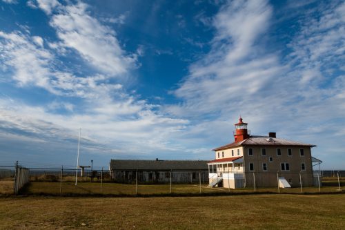 A lighthouse with a red top stands beside a weathered building under a blue sky with scattered clouds.