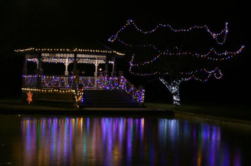 A beautifully lit gazebo surrounded by colorful holiday lights, reflecting in a calm pond at night.