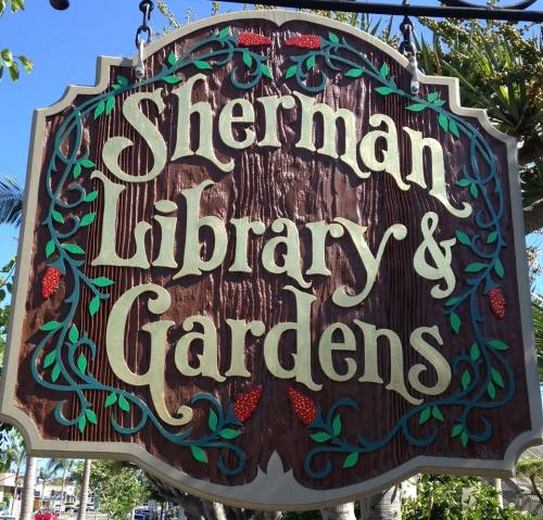 Sign for Sherman Library & Gardens, featuring decorative lettering and floral accents against a wooden background.