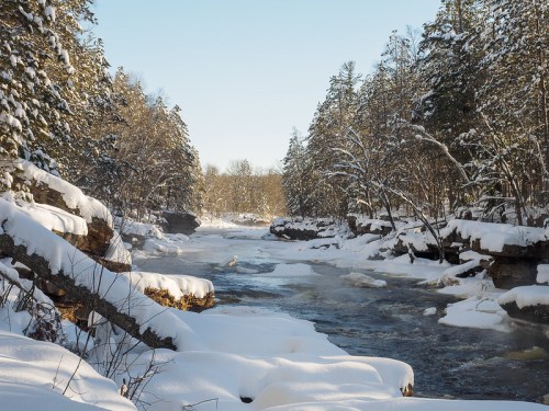 A serene winter landscape featuring a snow-covered river surrounded by trees and rocky banks under a clear blue sky.