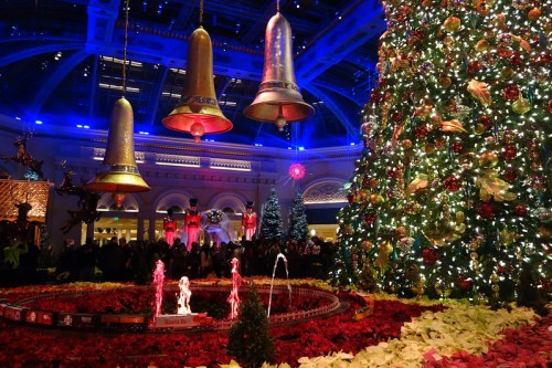 A festive scene featuring a large Christmas tree, decorative bells, and colorful lights, surrounded by poinsettias and a crowd.