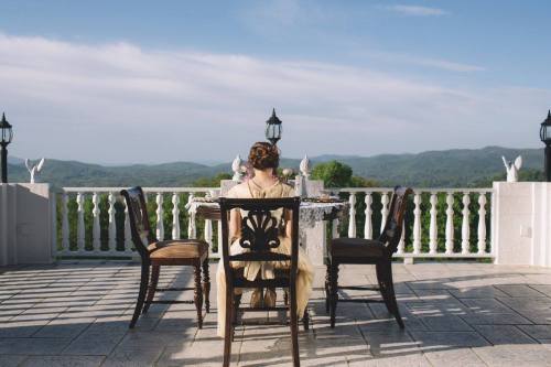 A woman in a vintage dress sits at a table on a balcony, overlooking a scenic mountain view.