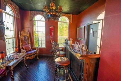 A cozy room with a bar, ornate seating, large windows, and a chess set, featuring warm red walls and elegant decor.