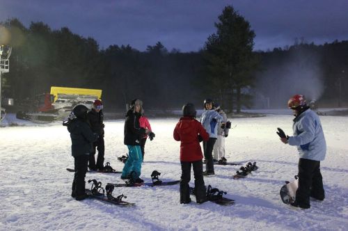 A group of six people in winter gear stands on snowboards, discussing in a snowy landscape at dusk.