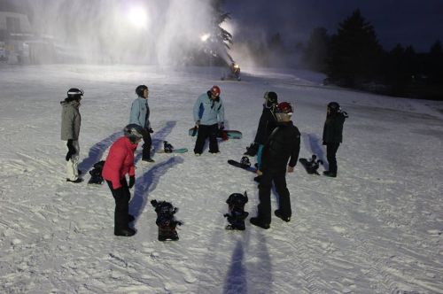 A group of snowboarders gathers on a snowy slope, preparing their gear under evening lights.