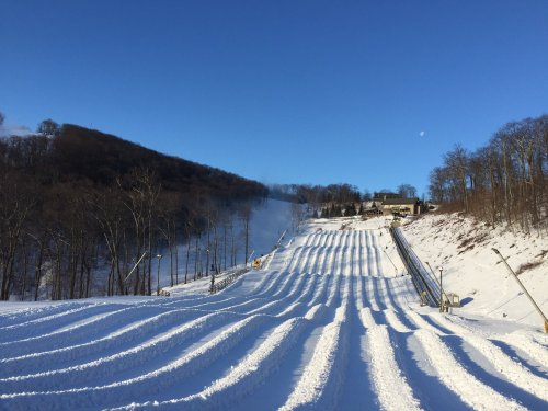 A snowy ski slope with groomed trails under a clear blue sky, surrounded by trees and a distant building.