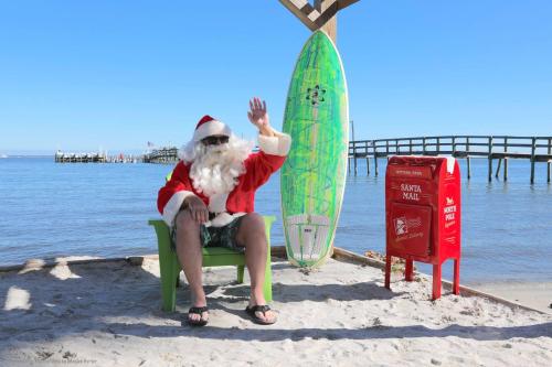 Santa in a beach chair waves, wearing sunglasses and a red suit, next to a surfboard and a mailbox labeled "Santa Mail."
