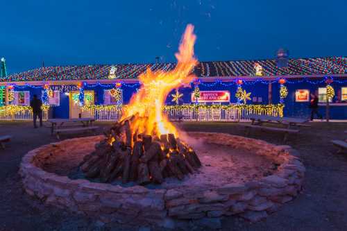 A cozy bonfire surrounded by stone, with a brightly lit building decorated for the holidays in the background.