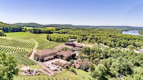 Aerial view of a vineyard estate surrounded by lush green fields and hills, with a river in the background.