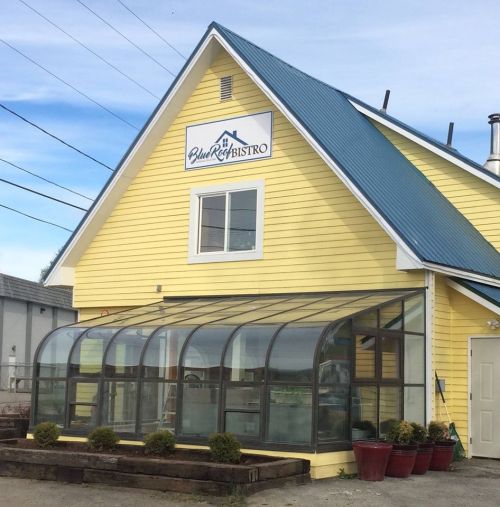 A yellow building with a blue roof and a glass greenhouse entrance, featuring a sign that reads "Blue Roof Bistro."
