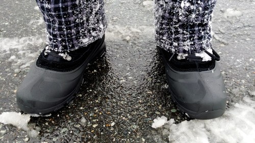 Close-up of waterproof boots on snowy ground, with snowflakes on dark plaid pants.