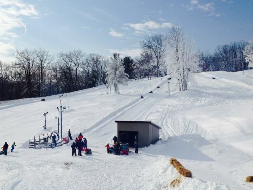 A snowy hill with people sledding and a small building at the base, surrounded by trees and a clear blue sky.