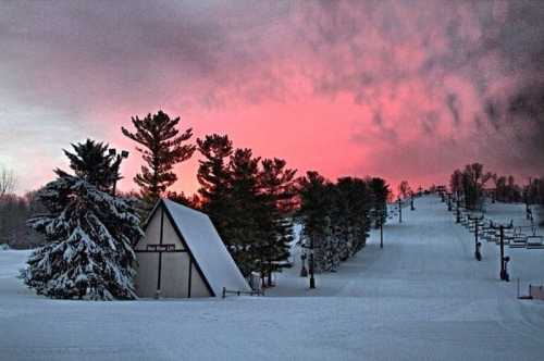 A snowy landscape at sunset, featuring a ski lodge and trees against a vibrant pink and purple sky.