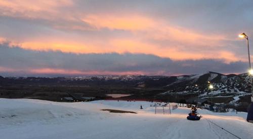 A snowy landscape at dusk, with colorful clouds and mountains in the background, and people tubing down a slope.