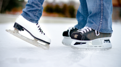 Close-up of two pairs of ice skates on an ice rink, one black and one white, with jeans visible.