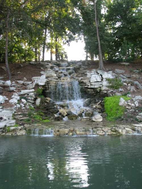 A serene waterfall cascades over rocks into a calm pond, surrounded by lush greenery and trees.