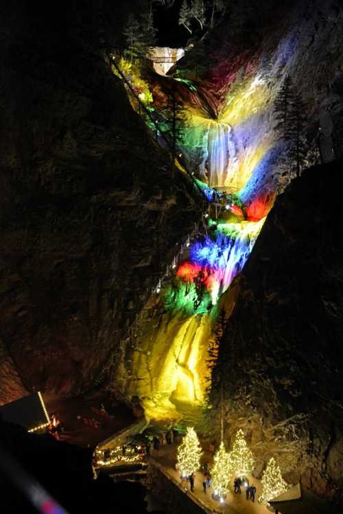 A colorful waterfall illuminated at night, surrounded by trees and festive decorations, with people gathered below.