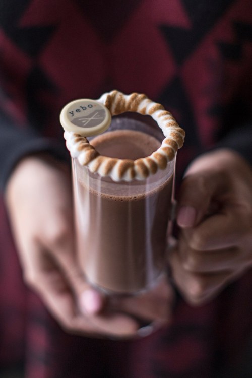 A person holding a glass of hot chocolate topped with a decorative whipped cream rim and a small round tag.