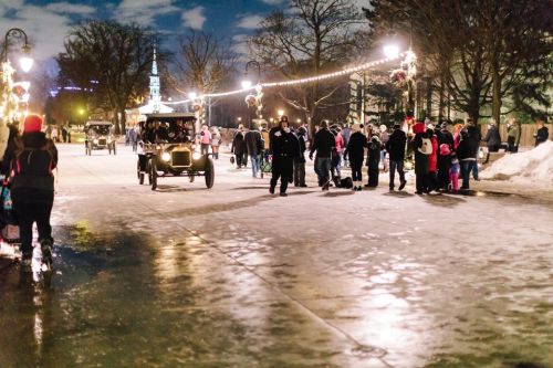 A winter evening scene with people walking on a snowy path, vintage cars, and festive lights in a park.