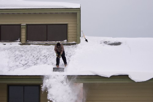 A person shovels snow off a roof, with a shovel nearby and a thick layer of snow covering the surface.