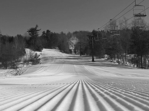 A snow-covered ski slope with freshly groomed tracks, surrounded by trees and ski lift towers in the background.