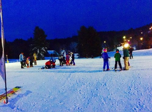 A snowy landscape at dusk with people skiing and gathering, surrounded by trees and lights in the background.