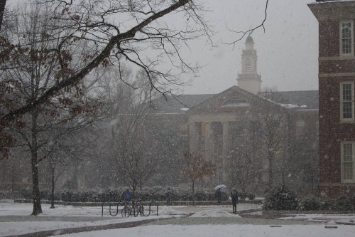 A snowy campus scene with trees, a building in the background, and a few people walking under umbrellas.