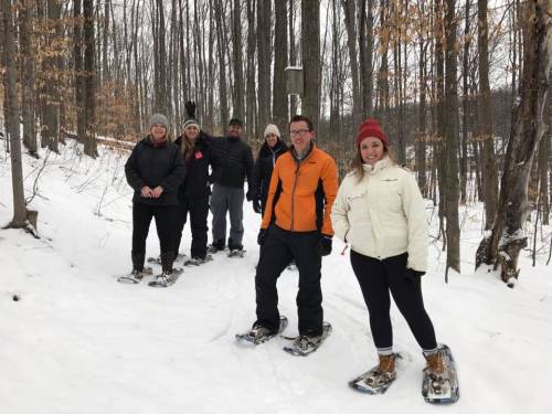A group of six people wearing winter clothing stand on a snowy trail in a forest, smiling and snowshoeing.