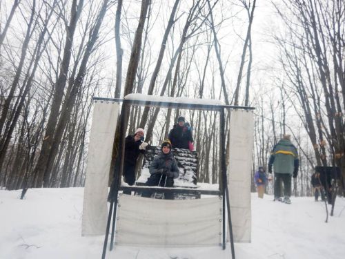 A group of people playfully poses in a snowy forest, with trees in the background and a snowy structure in the foreground.