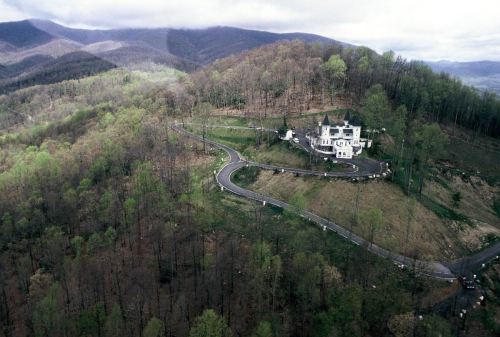 Aerial view of a white house on a hillside, surrounded by green trees and mountains under a cloudy sky.