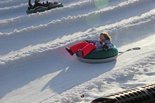 A child in a green snow tube slides down a snowy slope, wearing bright red pants and enjoying the winter fun.