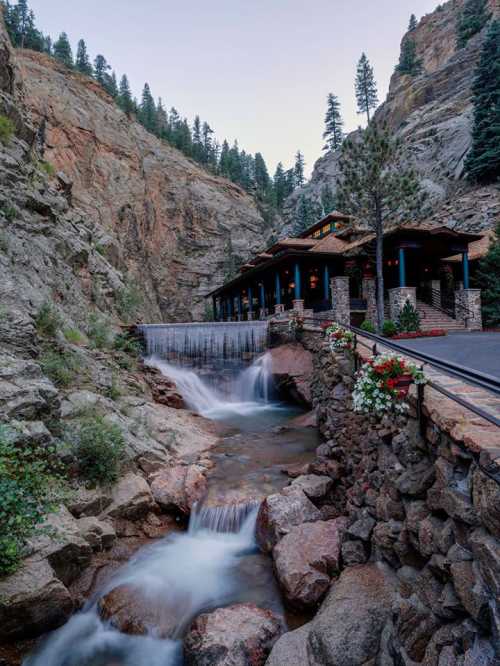 A serene waterfall flows beside a stone building nestled in a rocky landscape, surrounded by tall trees.