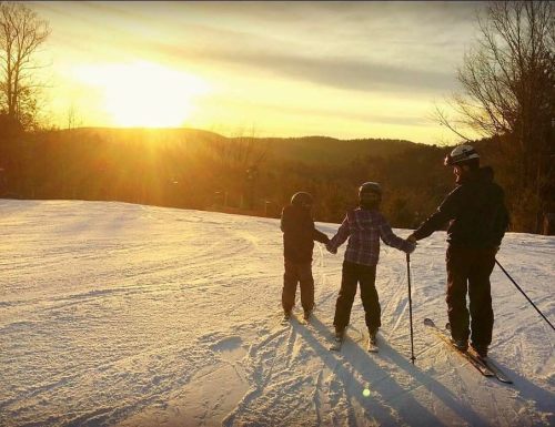 A parent and two children hold hands on a snowy slope at sunset, with mountains in the background.