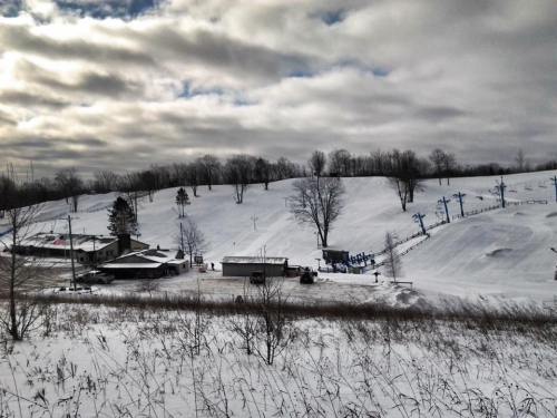 A snowy landscape with hills, trees, and ski lifts under a cloudy sky, featuring a small building in the foreground.