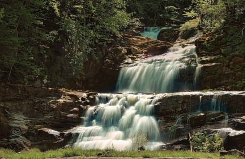 A serene waterfall cascading over rocky ledges, surrounded by lush green trees and soft sunlight.