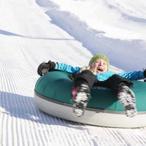 A child joyfully sliding down a snowy hill on a green snow tube, wearing winter clothing and gloves.