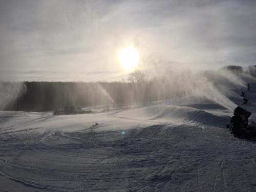 Snowmaking machines create mist in a snowy landscape under a bright sun, with trees in the background.