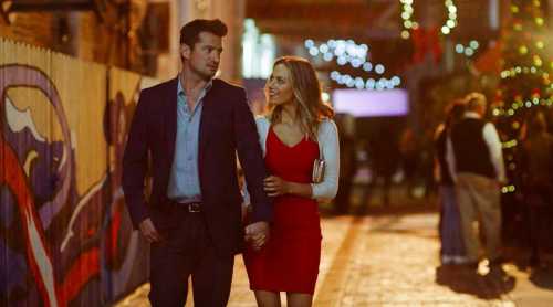 A couple walks hand in hand down a festive street, surrounded by holiday lights and decorations.