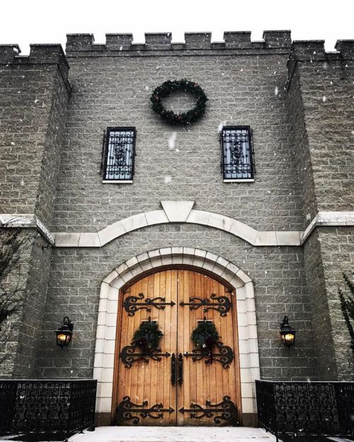A stone castle entrance adorned with wreaths, featuring wooden doors and snow gently falling.
