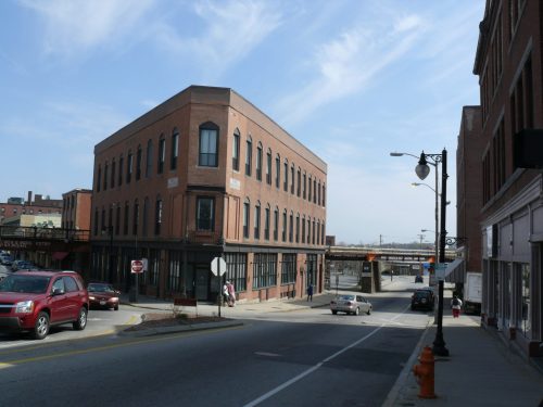 A brick building on a street corner, with a clear sky and a bridge in the background. Cars parked along the road.