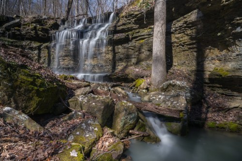 A serene waterfall cascades over rocky cliffs, surrounded by moss-covered stones and bare trees in a tranquil forest setting.