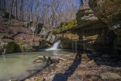 A serene waterfall cascades into a calm pool, surrounded by rocky cliffs and bare trees under a clear blue sky.